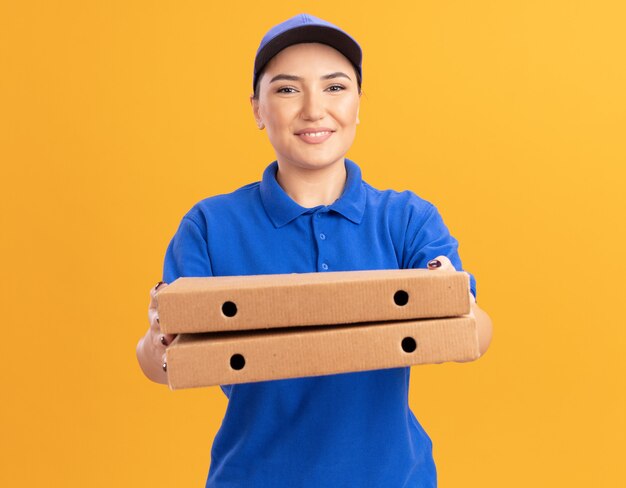 Happy young delivery woman in blue uniform and cap holding pizza boxes looking at front smiling confident standing over orange wall