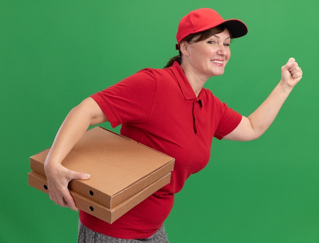 Free Photo happy young delivery woman in red uniform and cap rush running for delivering pizza boxes for customer over green wall