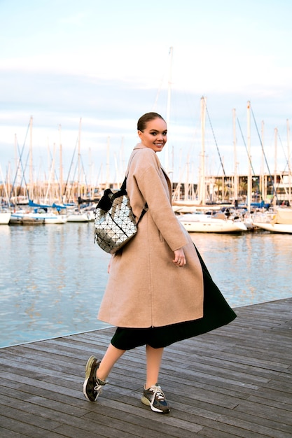 Free Photo happy young elegant woman walking at luxury yacht club of barcelona, wearing coat sneakers and backpack, mid season touristic time .