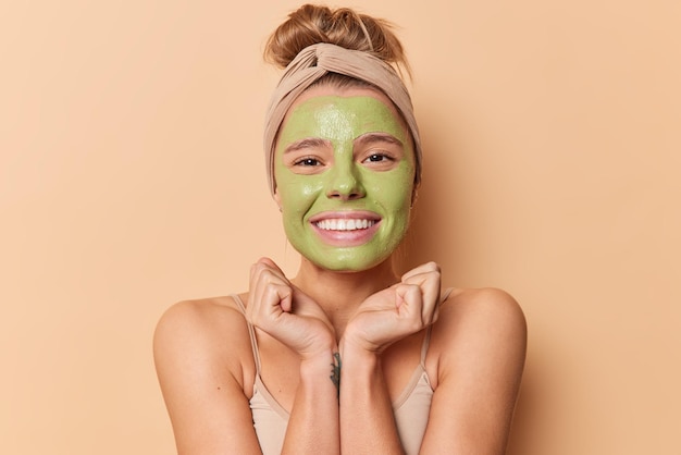 Free photo happy young european woman keeps hands under chin smiles gladfully applies green nourishing mask wears headband undergoes beauty procedures for skin treatment isolated over beige background.