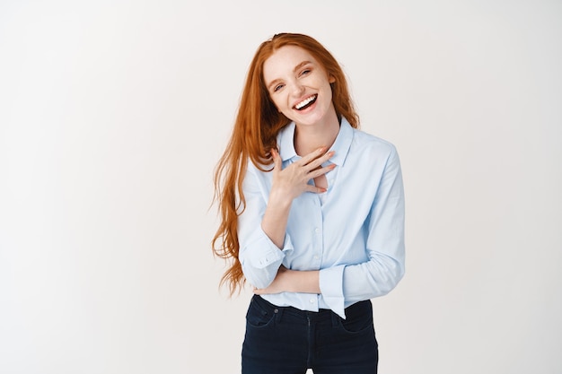 Free Photo happy young female with long red hair pointing at herself, laughing and looking confident, standing over white wall