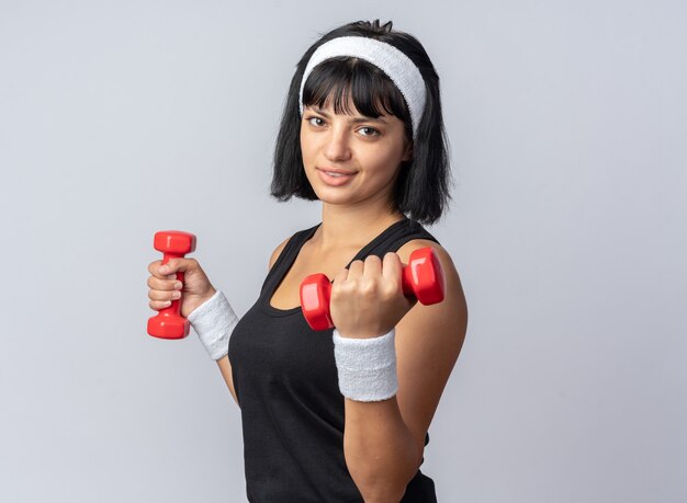 Happy young fitness girl wearing headband holding dumbbells doing exercises looking confident standing over white background