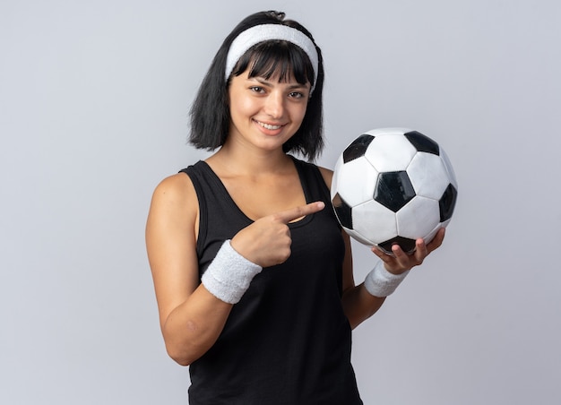 Happy young fitness girl wearing headband holding soccer ball pointing with index finger at it smiling cheerfully looking at camera standing over white background