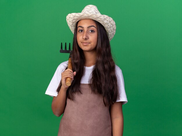 Happy young gardener girl in apron and summer hat holding mini rake  with smile on face standing over green wall