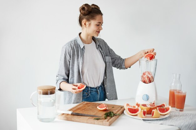 Happy young girl blending healthy detox fresh grapefruit smoothie over white wall.