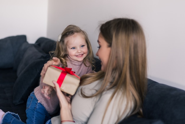 Happy young girl surprising her mother with gift at home in living room