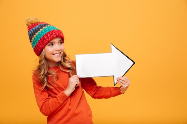 Happy Young girl in sweater and hat looking and pointing with paper arrow away over orange