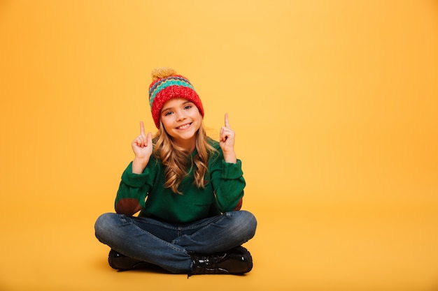 Happy Young girl in sweater and hat sitting on the floor while pointing up and looking at the camera over orange