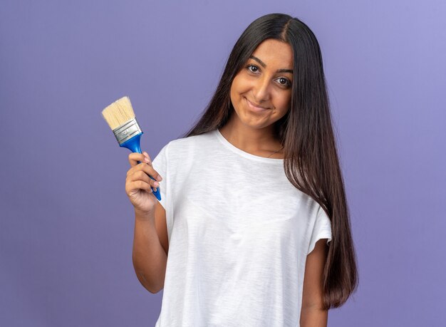 Happy young girl in white t-shirt holding paint brush looking at camera with smile on face standing over blue background