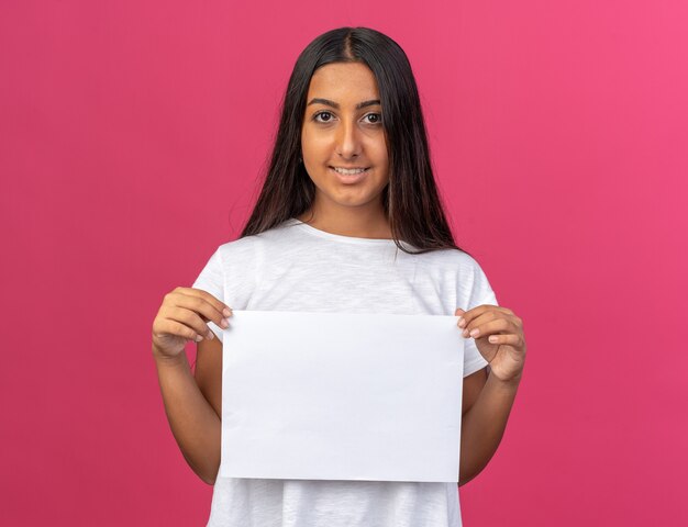 Happy young girl in white t-shirt holding white blank sheet of paper looking at camera smiling cheerfully 