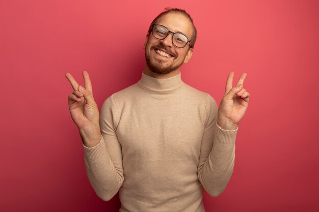 Happy young handsome man in beige turtleneck and glasses looking at front smiling cheerfully showing v-sign with both hands standing over pink wall