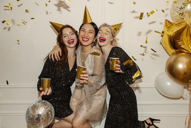 Happy young interracial ladies in dresses are walking at party with booze balloons and confetti on white background Happy weekend concept