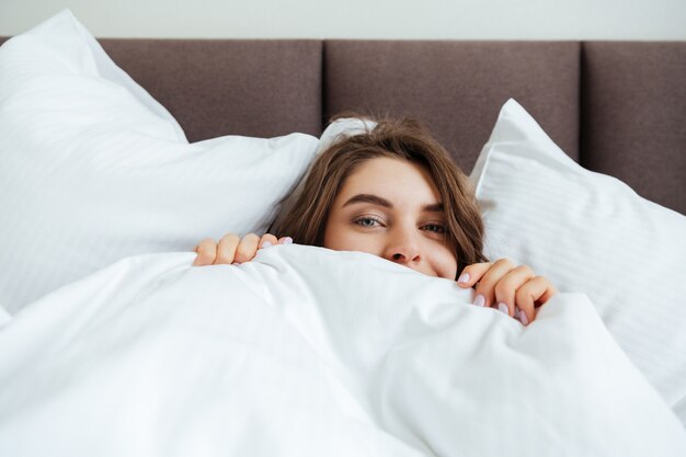 Happy young lady lies in bed at home under a blanket.