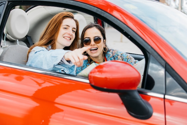 Happy young lady pointing to cheerful woman and sitting in car