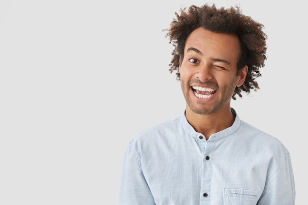 Happy young man blinks eye, has positive expression, has crisp dark hair, dressed casually