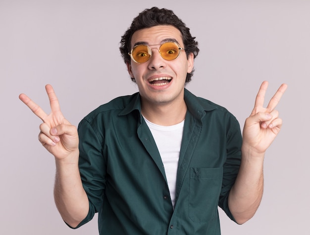 Happy young man in green shirt wearing glasses looking at front smiling cheerfully showing v-sign standing over white wall