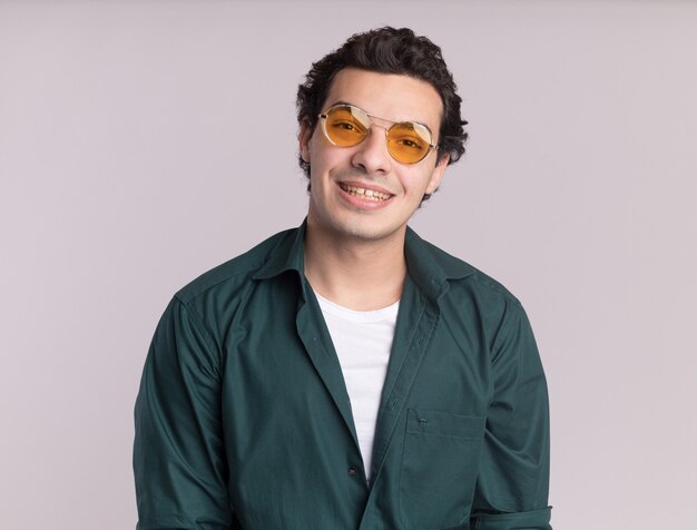 Happy young man in green shirt wearing glasses looking at front with smile on face standing over white wall