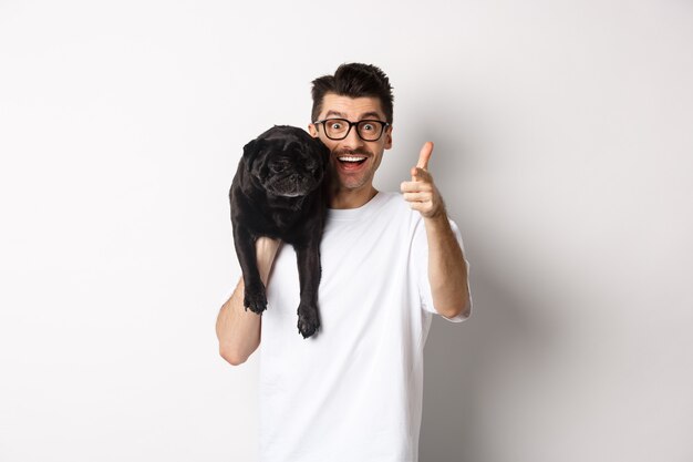 Happy young man holding cute black dog on shoulder and pointing at camera. Hipster guy carry pug on shoulder and staring at camera excited, standing over white background.