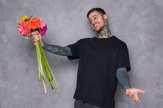 Free photo happy young man holding gerbera flowers in hand shrugging against grey wall