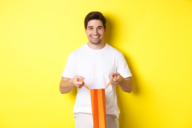 Free photo happy young man open shopping bag with present, smiling at camera, standing against yellow background.