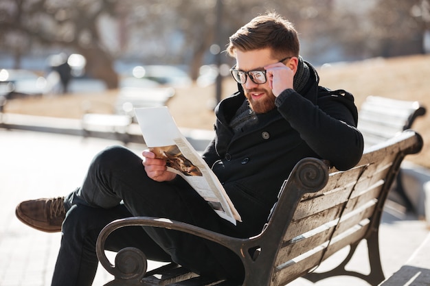 Happy young man reading newspaper on bench in the city