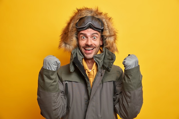 Free photo happy young man rejoices winter came raises clenched fists wears gloves and warm jacket with hood