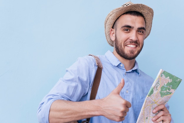 Free Photo happy young man showing thumb up gesture and holding map against blue background