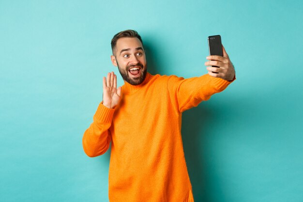 Happy young man video calling, talking online with mobile phone, saying hello to smartphone camera and waving hand friendly, standing over light turquoise wall.