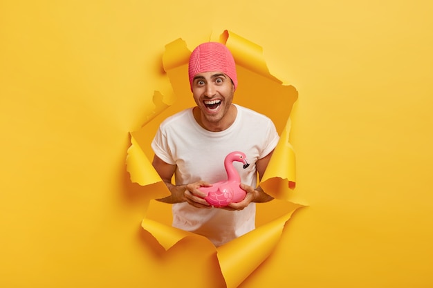 Happy young man wears rubber pink swimcap, wears white t shirt, holds swim ring in shape of flamingo