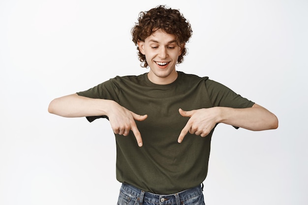 Happy young man with curly hair pointing and looking down smiling and laughing reading logo advertisement standing over white background