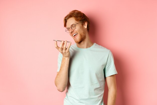 Happy young man with red hair smiling pleased while record voice message on smartphone talking to vi...