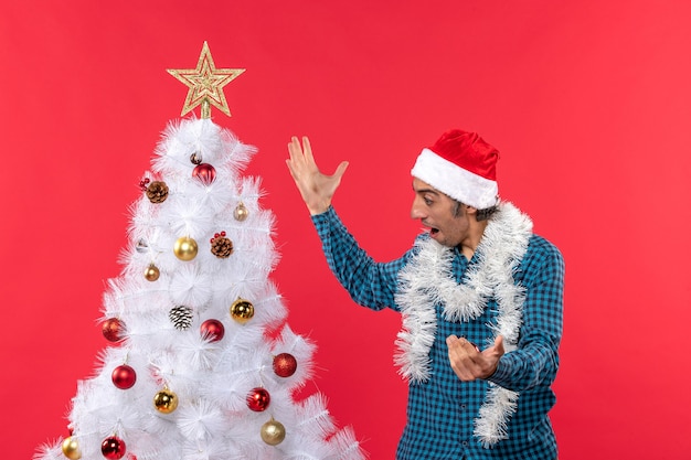 Free photo happy young man with santa claus hat in a blue stripped shirt and looking at xsmas tree suprisingly red