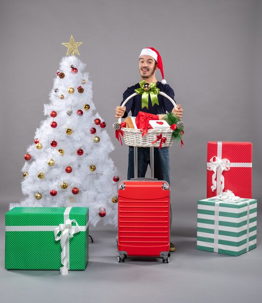 happy young man with santa hat holding gift basket with both hands near white xmas tree on grey