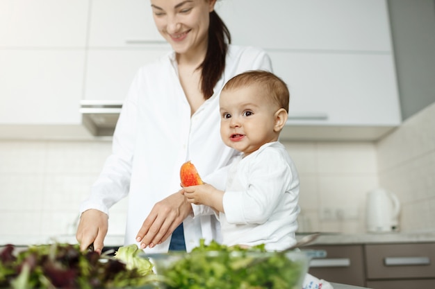 Happy young mother cooking breakfast in light kitchen with her little cute son. Kid eating peach with funny expression while mom works.