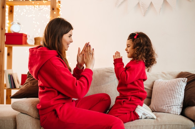 Happy young mother playing with daughter on sofa. Indoor shot of charming mom and preteen kid in red attire.