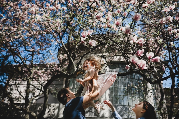 Free photo happy young parents with a little daughter stand under blooming pink tree outside