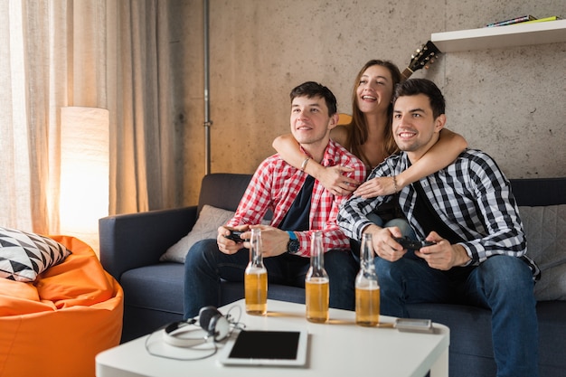 Happy young people playing video games, having fun, friends party at home, hipster company together, two men one woman, smiling, positive, relaxed, emotional, laughing, competition