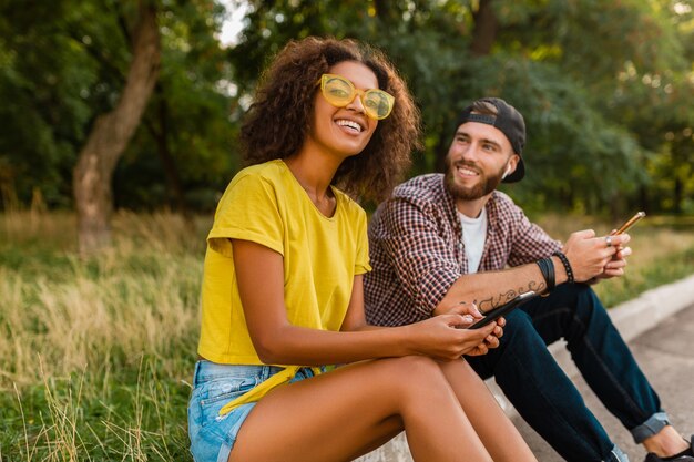 Happy young smiling friends sitting park using smartphones, man and woman having fun together