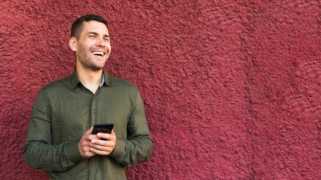 Happy young stubble man holding cellphone standing near rough wall