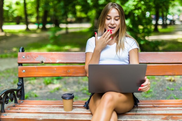 Happy young student with a tablet sitting on the bench and reading in a summer park.