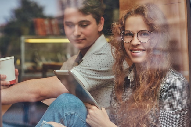 Happy young students drinking coffee and using a digital tablet sitting on a window sill at a college campus during a break.