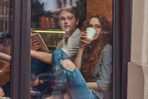 Happy young students drinking coffee and using a digital tablet sitting on a window sill at a college campus during a break.