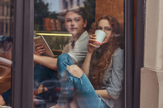 Free photo happy young students drinking coffee and using a digital tablet sitting on a window sill at a college campus during a break.