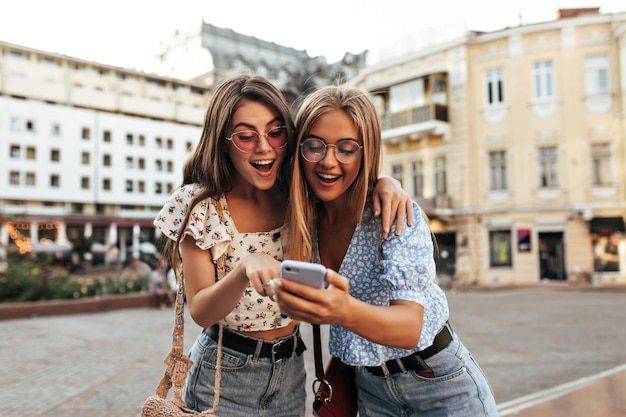 Happy young tanned girls in colorful sunglasses and stylish cropped blouses look at phone screen and laugh Blonde and brunette women read message