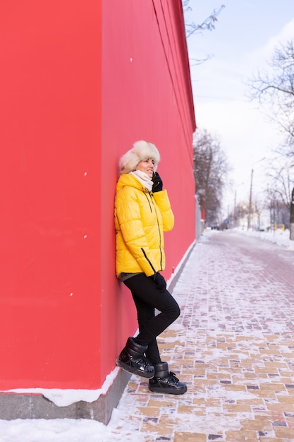 Free photo happy young woman on a background of a red wall in warm clothes on a winter sunny day smiling and talking on the phone on a snowy city sidewalk