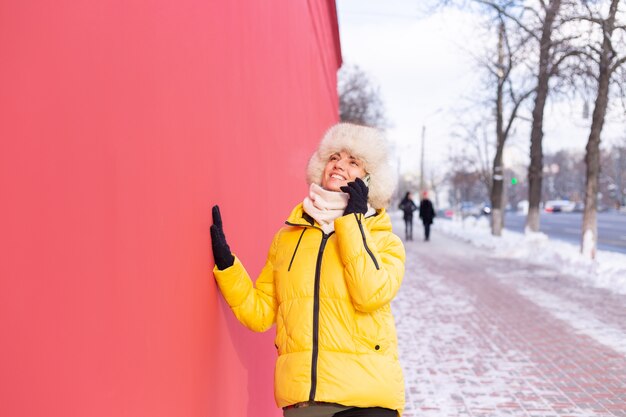 Happy young woman on a background of a red wall in warm clothes on a winter sunny day smiling and talking on the phone on a snowy city sidewalk