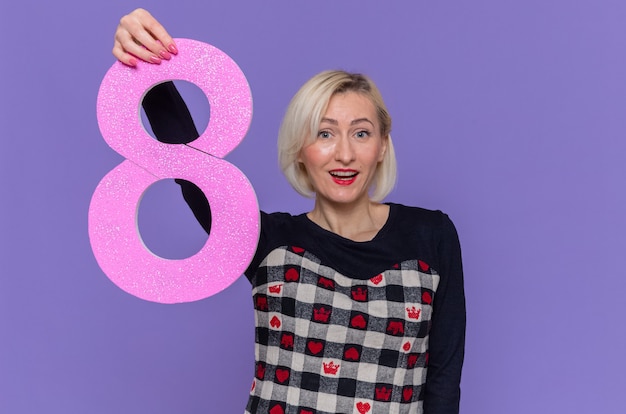 Free Photo happy young woman in beautiful dress holding number eight made from cardboard looking at front smiling cheerfully celebrating international women's day standing over purple wall