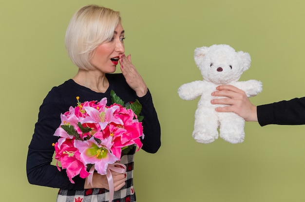 Free photo happy young woman in beautiful dress with bouquet of flowers looking surprised smiling while receiving teddy bear as a gift celebrating international women's day standing over green wall