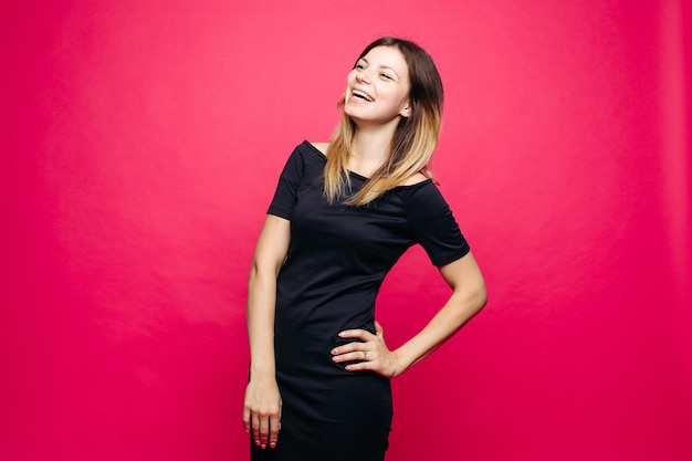 Happy young woman in black dress posing at studio and lauthing looking away Positivity emotionaly girl holding hand on waist standing against pink background Concept of shopping fashionbeauty