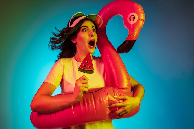 Free Photo happy young woman in a cap and in a rubber flamingo with the candy on trendy blue neon